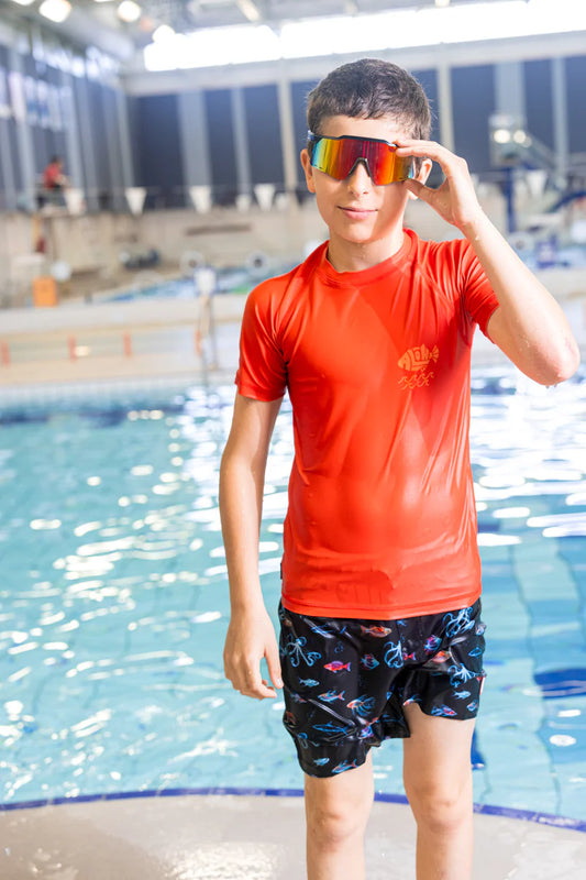 Garçon portant Lunette de soleil - Nanö à verre miroir rouge, t-shirt sportif orange, short noir motifs poissons, vue de face.