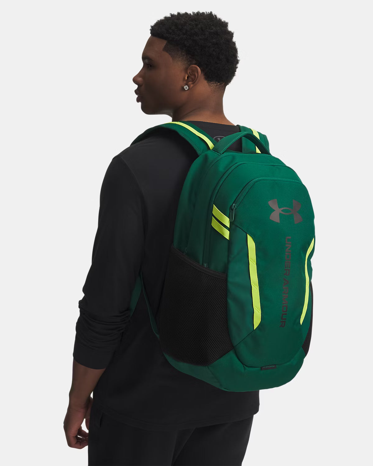 Sac à dos - Under Armour vert foncé avec détails jaunes fluo, en tissu résistant, porté sur le dos par un jeune homme, vue de profil arrière.