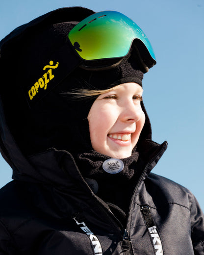 Cagoule - Deux Par Deux noire en tissu technique, portée par enfant, vue de profil avec lunettes de ski vertes réfléchissantes