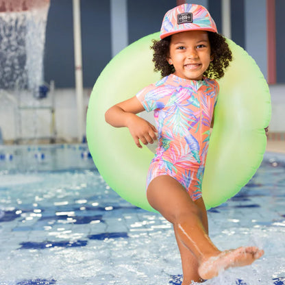 Maillot de bain - Nanö, une pièce manches courtes rose à motifs tropicaux bleus et verts, fille, porté au bord de piscine, vue de face.