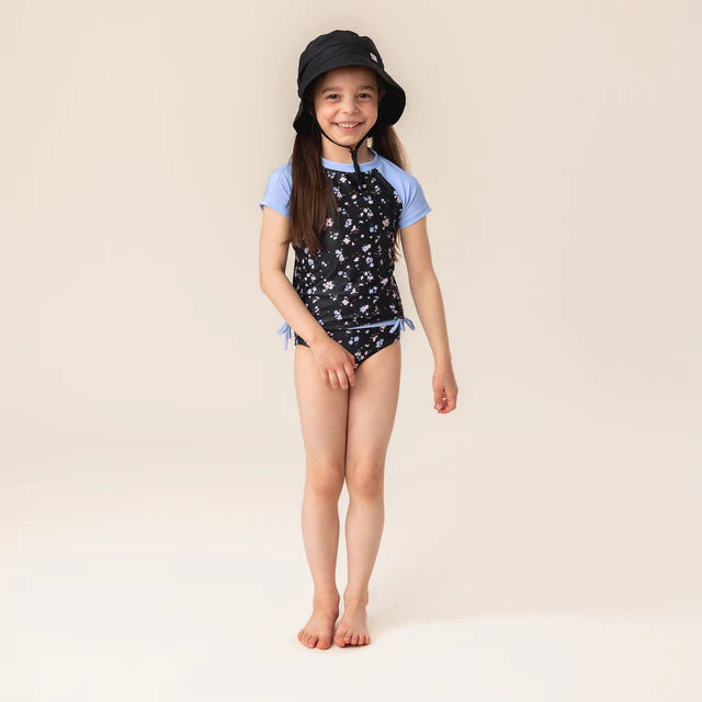 Maillot de bain - Nanö, deux pièces noir à motifs floraux blancs et bleus, manches courtes bleu clair, pour enfant fille, porté de face.