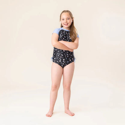 Maillot de bain - Nanö à manches courtes bleu et noir avec motifs floraux blancs, pour fille, porté de face sur mannequin enfant.