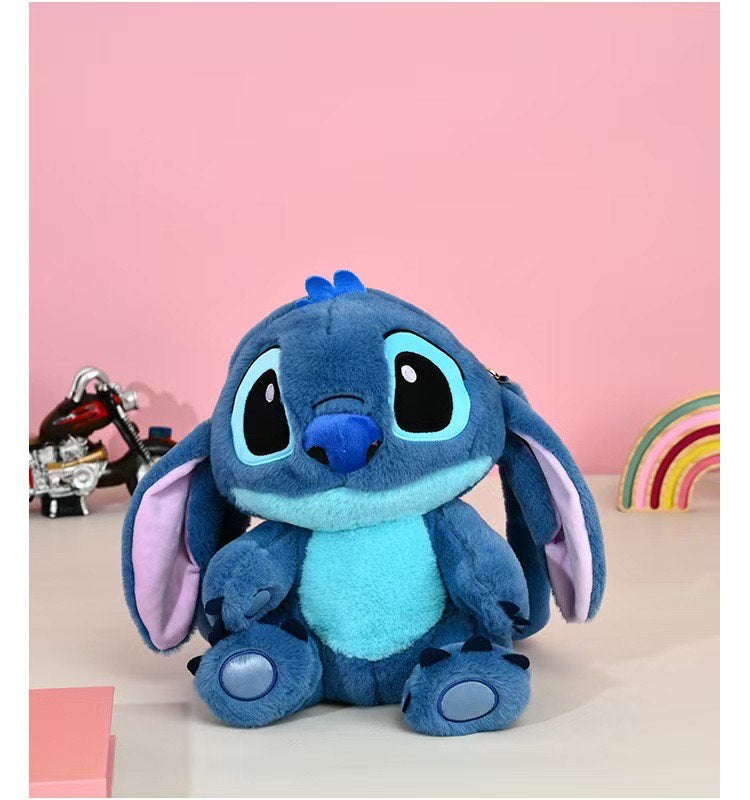 Une peluche Stitch bleue avec de grandes oreilles flottantes est assise à côté d'un TOMMY Mini Sac à Dos - Stitch Bleu sur une surface blanche. Un petit jouet moto et une décoration arc-en-ciel apparaissent sur un fond rose.