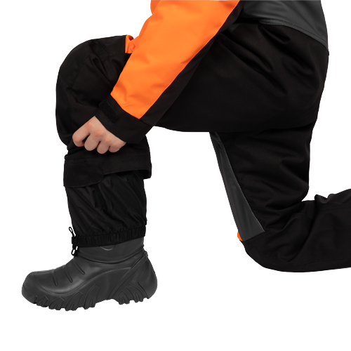 Habit de neige 1 pièce - FXR noir et orange, détail porté en position accroupie avec botte d’hiver noire