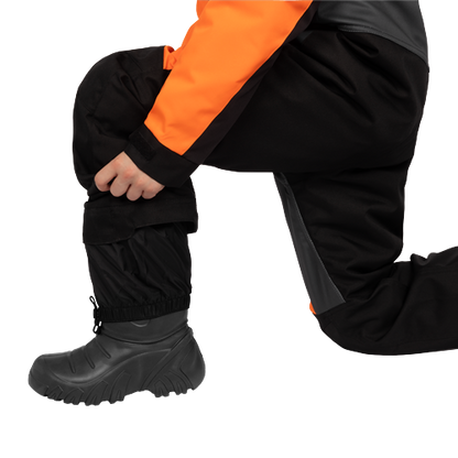 Habit de neige 1 pièce - FXR noir et orange, détail porté en position accroupie avec botte d’hiver noire